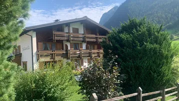 Traditionelles Gebäude im alpinen Stil mit Holzbalkonen, umgeben von grünen Büschen und Bergblick.
