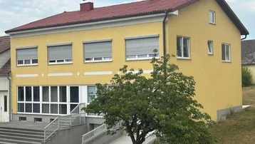 Gelbes Gebäude mit rotem Satteldach, vielen Fenstern und Rollläden, umgeben von Grünflächen.