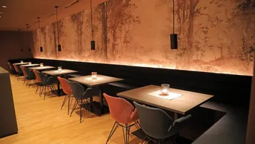 Gemütlicher Restaurantbereich mit Holzboden, gepolsterten Bänken und einer Wandtapete mit Waldmotiv, beleuchtet von Pendelleuchten.