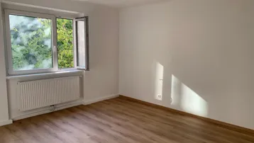 Helles Zimmer mit Laminatboden und geöffnetem Fenster, das einen Blick ins Grüne bietet.