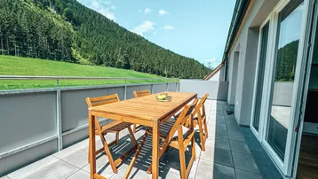 Große Terrasse mit Holztisch und Stühlen, umgeben von grünen Bergen unter blauem Himmel.
