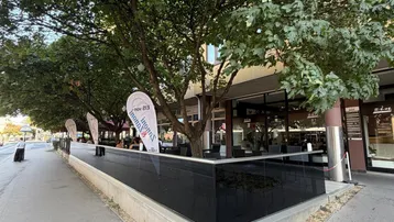 Außenbereich eines Cafés mit schattenspendenden Bäumen und Sitzgelegenheiten entlang der Straße.