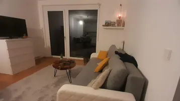 Gemütliches Wohnzimmer mit Sofa, Fernseher und Zugang zu einem Balkon oder einer Terrasse.