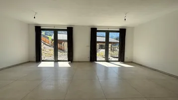Helles, leeres Wohnzimmer mit großen Fenstertüren und Blick auf die umliegende Landschaft.
