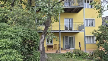 Gelbe Fassade eines Mehrfamilienhauses mit Balkonen und einer möblierten Terrasse im Erdgeschoss.