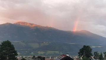 Malerischer Bergblick mit einem Regenbogen über den Gipfeln und Dächern im Vordergrund.