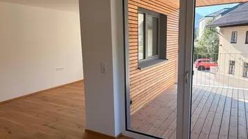 Helles Zimmer mit Holzboden und Zugang zu einem Balkon mit Holzverkleidung.