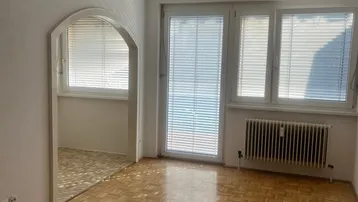 Helles Zimmer mit Parkettboden, großen Fenstern und Heizkörper, verbunden durch einen Rundbogen.