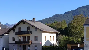 Traditionelles Haus mit Satteldach und Balkonen, umgeben von Bergen und grüner Landschaft.