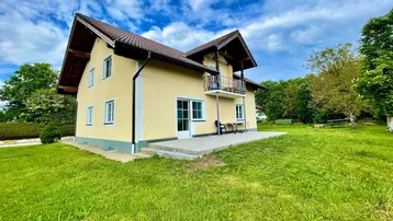 Gelbes Einfamilienhaus mit braunem Dach, umgeben von grünem Rasen und Bäumen unter blauem Himmel.