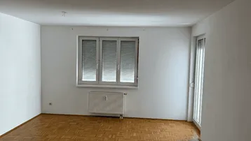 Heller, leerer Raum mit Parkettboden, Fenster mit Rollläden und Zugang zum Balkon.