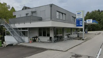 Gebäude mit moderner Fassade, Außentreppe und einer Terrasse mit Sitzgelegenheiten neben einer Tankstelle.