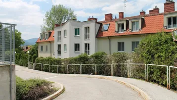 Mehrere Gebäude mit roten Ziegeldächern und grünen Hecken entlang einer kurvigen Straße.
