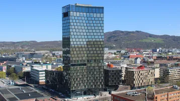 Hohes modernes Gebäude mit Glasfassade, umgeben von städtischer Bebauung und grünen Hügeln im Hintergrund.