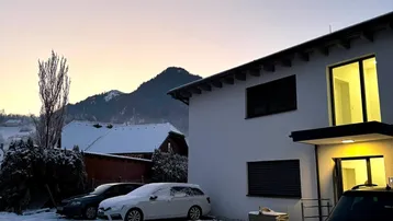 Modernes Haus in verschneiter Landschaft mit Bergen im Hintergrund bei Sonnenaufgang.