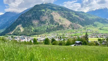 Panoramablick auf ein grünes Tal mit einem Dorf, umgeben von Bergen unter blauem Himmel.