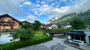 Blick auf eine Wohnsiedlung mit traditionellen Häusern, grünen Gärten und Bergen im Hintergrund unter bewölktem Himmel.