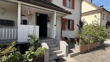 Vorderansicht eines Hauses mit hellen Wänden, roten Fensterläden und einer kleinen Treppe zum Eingang.