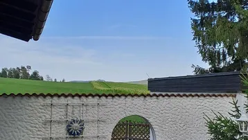 Blick über eine weiße Mauer mit Torbogen auf grüne Felder und den blauen Himmel.