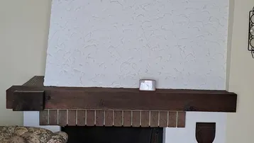 Traditioneller Kamin mit weißer Strukturputzoberfläche und Holzbalken, neben einem Sessel.