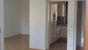 Blick von einem leeren Raum in eine helle Küche mit modernen Einbauschränken und Geräten.