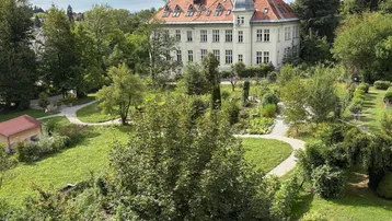 Großes klassisches Gebäude mit rotem Dach, umgeben von weitläufigen grünen Gärten und Bäumen unter bewölktem Himmel.