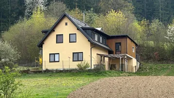 Gelbes Einfamilienhaus mit schwarzem Dach, umgeben von grünen Hügeln und Wald.