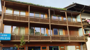 Mehrstöckiges Gebäude mit Holzfassade und mehreren Balkonen, umgeben von grüner Bepflanzung auf dem Dach.