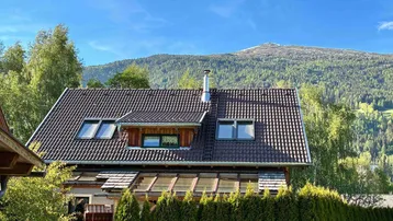Traditionelles Haus mit Satteldach und Dachfenstern, umgeben von grüner Hecke und Bergen.