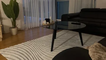 Gemütliches Wohnzimmer mit dunklem Ledersofa, modernem Glastisch und großem Fenster mit Vorhängen.