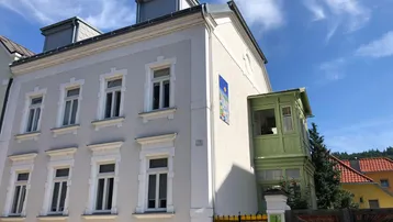 Historisches Gebäude mit heller Fassade, grünen Fensterläden und Dachgauben unter blauem Himmel.