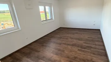 Leeres Zimmer mit Laminatboden und großen Fenstern, die einen Blick ins Grüne bieten.