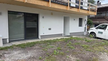 Eingangsbereich eines Gebäudes mit weißer Fassade und einem darüberliegenden Holzbalkon.