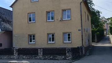 Gelbes Mehrfamilienhaus mit Satteldach an einer Straßenecke, umgeben von Bäumen und einem kleinen Zaun.