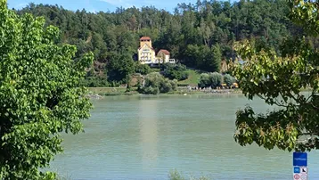 Panoramablick auf einen ruhigen See mit einem historischen Gebäude auf einem bewaldeten Hügel im Hintergrund.