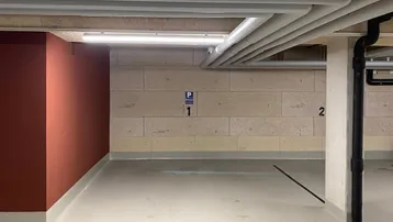 Helle Tiefgarage mit sichtbaren Deckenleitungen, Steinwänden und markierten Parkplätzen.