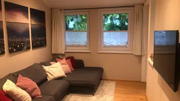 Gemütliches Wohnzimmer mit großem Ecksofa, Holzboden und Blick ins Grüne durch die Fenster.