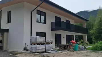 Modernes Haus im Bauzustand mit weißer Fassade, schwarzem Balkon und Blick auf die Berge.