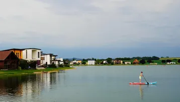 Seeufer mit modernen Häusern und einer Person beim Stand-Up-Paddling an einem bewölkten Tag.