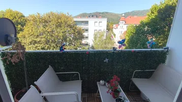 Möblierter Balkon mit Holzboden, grüner Sichtschutzwand und Blick auf die Stadt und Hügel.