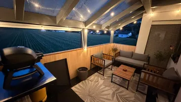 Überdachte Terrasse mit gemütlichen Sitzgelegenheiten, Grill und Blick auf weite Felder in der Abenddämmerung.