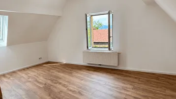 Heller Raum im Dachgeschoss mit Holzboden, schrägen Wänden und Ausblick ins Grüne.