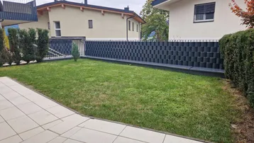 Gepflegter Gartenbereich mit Rasenfläche und gepflasterter Terrasse, umgeben von einer modernen Zaunanlage.