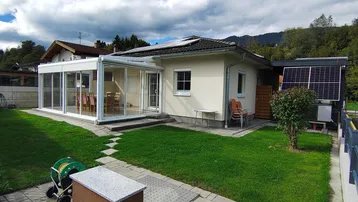 Modernes Haus mit Wintergarten, gepflegtem Rasen und Photovoltaikanlage vor bergiger Kulisse.