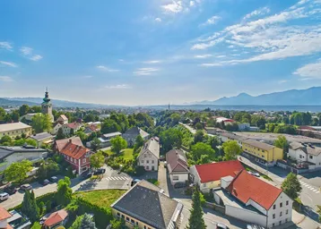 Panoramablick über eine Stadt mit Häusern, viel Grün und Bergen im Hintergrund unter blauem Himmel.