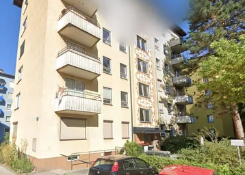 Mehrfamilienhaus mit mehreren Balkonen und Bäumen in der Umgebung, mit geparkten Autos davor.