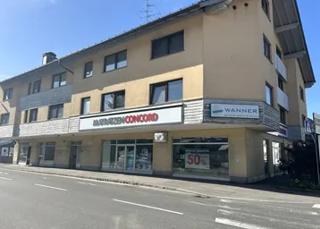 Gebäudeansicht mit Ladenlokalen im Erdgeschoss und Wohnungen darüber an einer Straße gelegen.