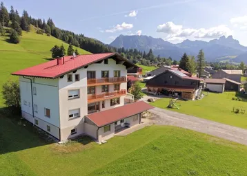 Großes Wohnhaus mit rotem Satteldach und Balkonen, umgeben von grünen Wiesen und Bergen.