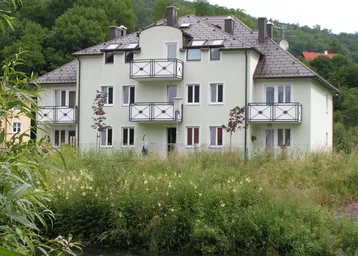 Mehrfamilienhaus mit hellgrüner Fassade, mehreren Balkonen und Blick auf die umliegende Natur und einen Fluss.