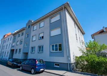 Mehrstöckiges Wohngebäude mit hellgrauer und blauer Fassade unter klarem blauem Himmel und parkenden Autos.